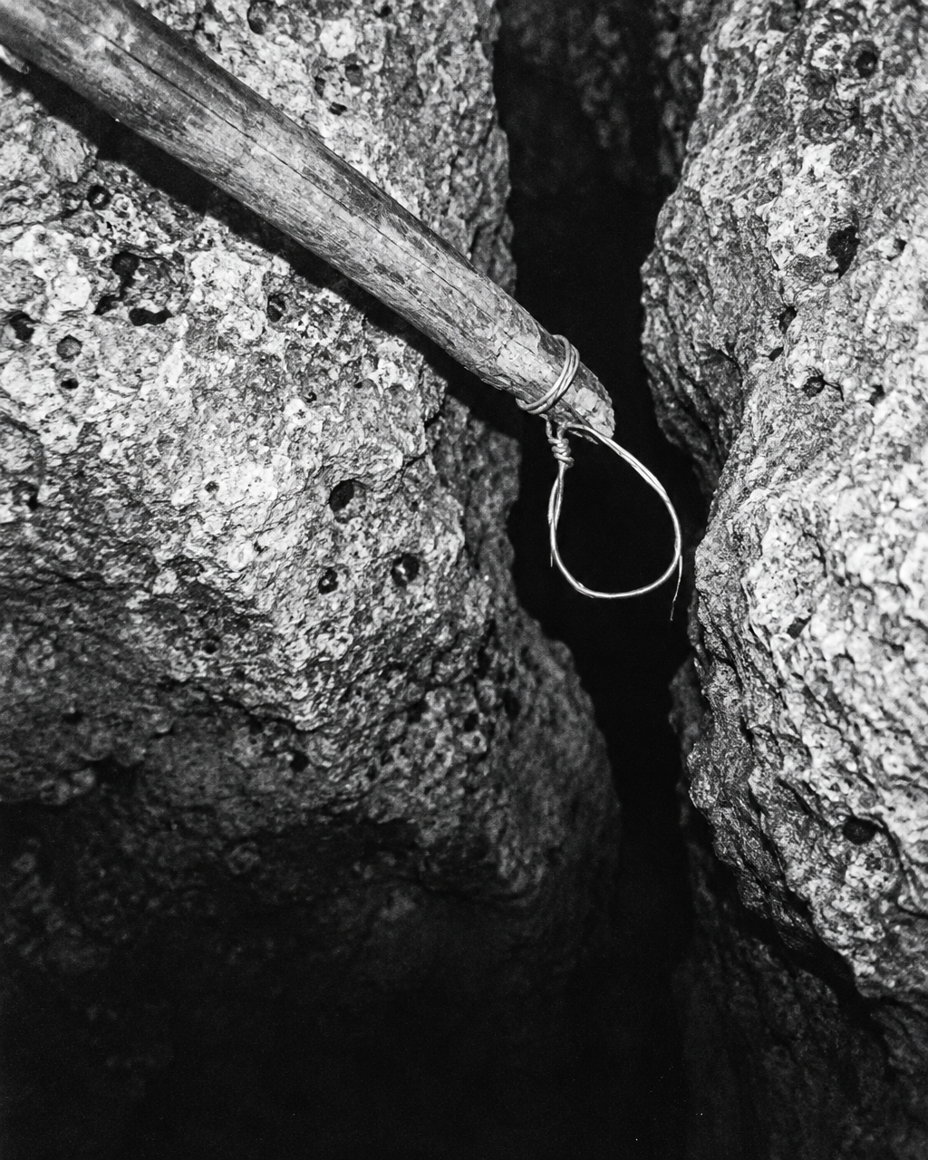 Fotografía en blanco y negro de estilo antiguo: un lazo de alambre en la punta de una vara de madera, junto a la boca de una grieta profunda en roca caliza