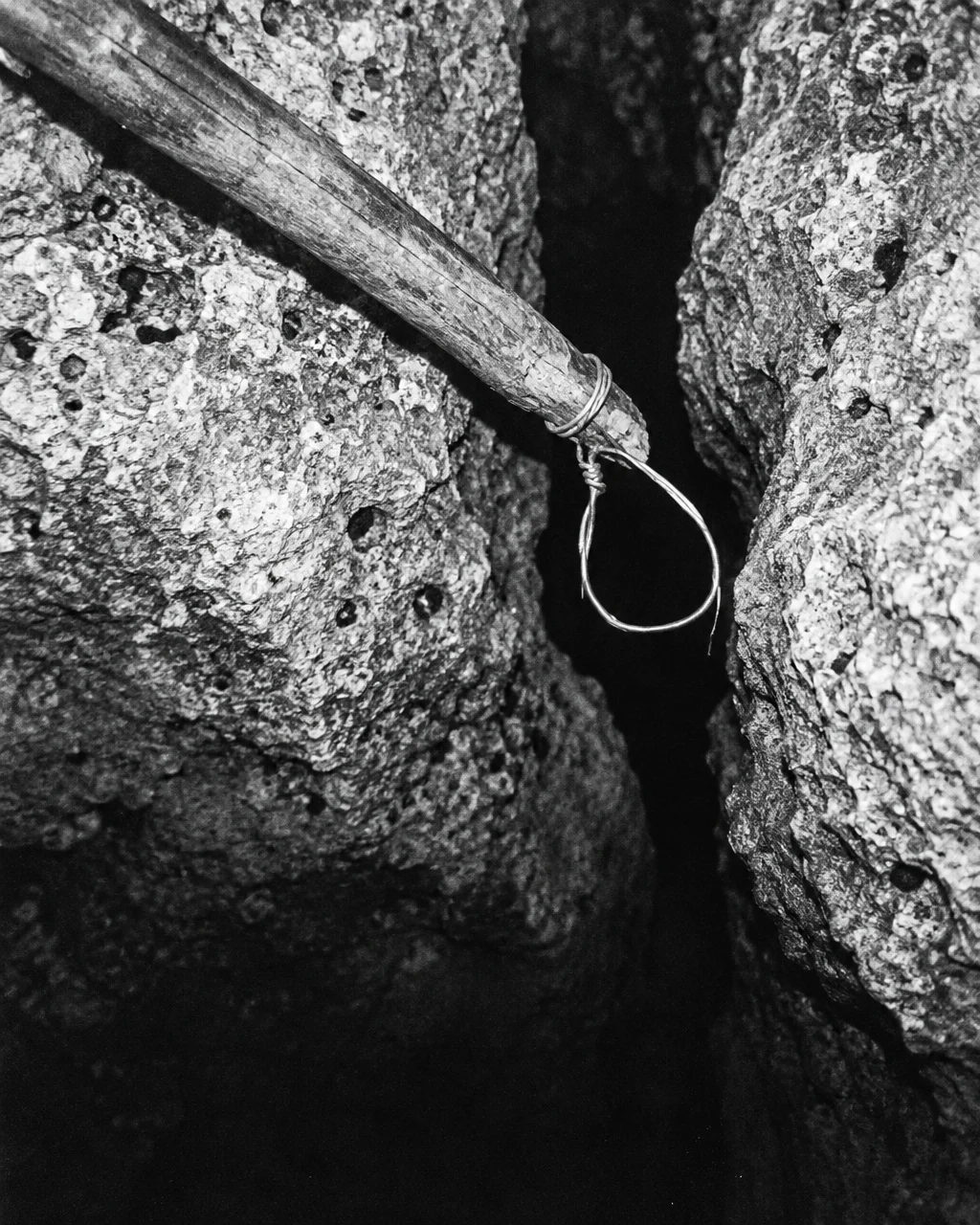 Fotografía en blanco y negro de estilo antiguo: un lazo de alambre en la punta de una vara de madera, junto a la boca de una grieta profunda en roca caliza
