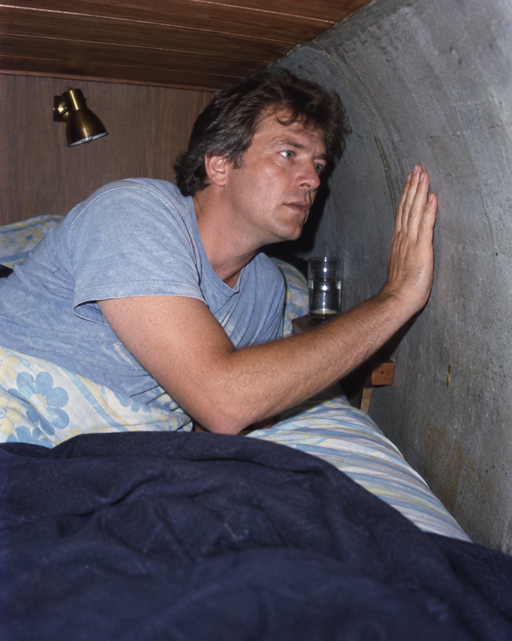 Persona de mediana edad sentada y despierta en una cama estrecha, dentro de un dormitorio pequeño en el casco de concreto de una casa flotante, iluminada por un flash directo.