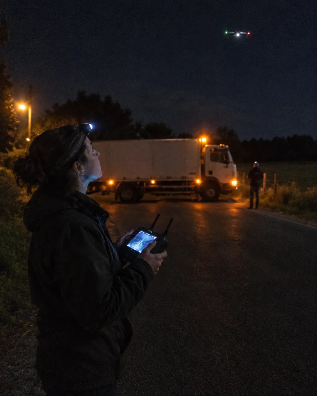 De noche, un trabajador con linterna en la cabeza maneja un dron junto a un camión atravesado en una carretera rural