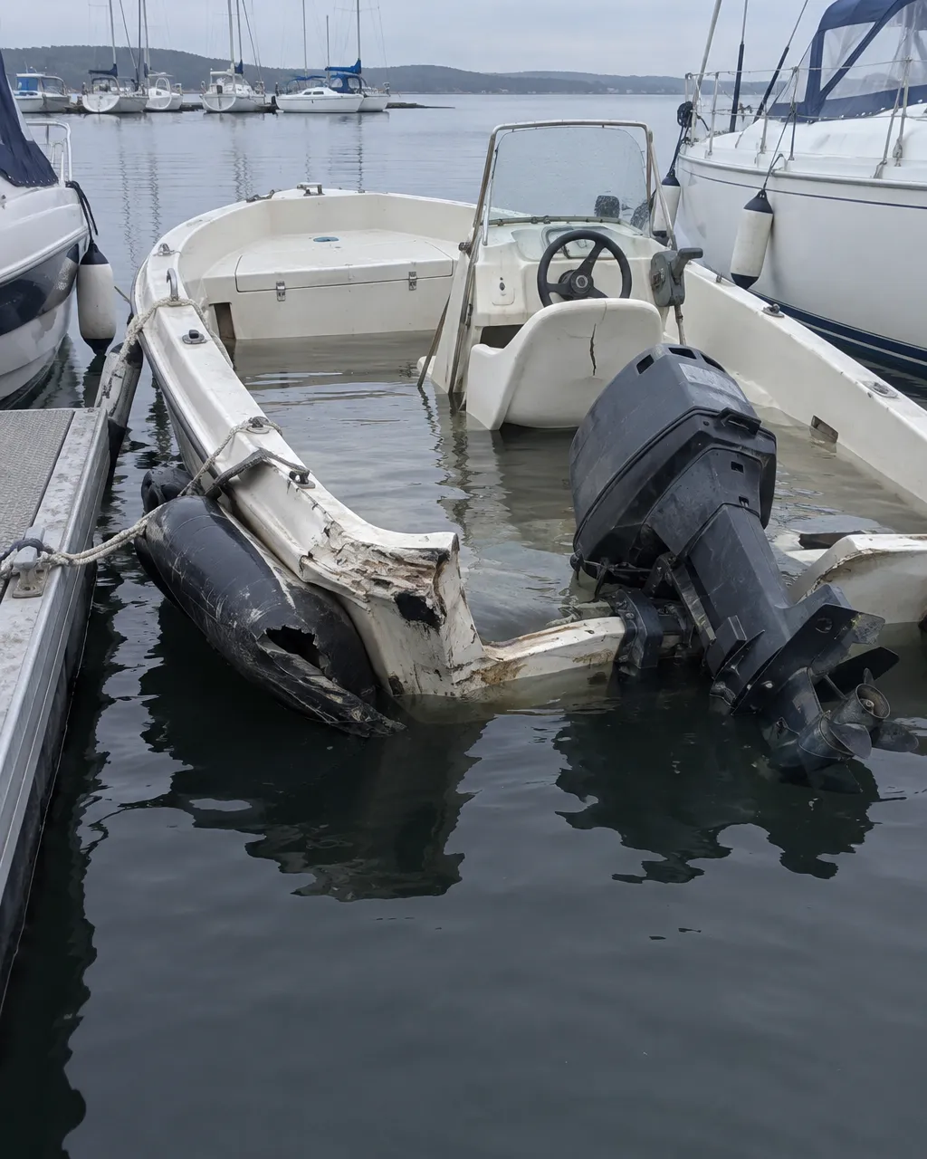 Un bote de motor pequeño amarrado a un pontón, con la popa hundida y parcialmente inundada, el motor fuera de borda torcido y una defensa de goma aplastada