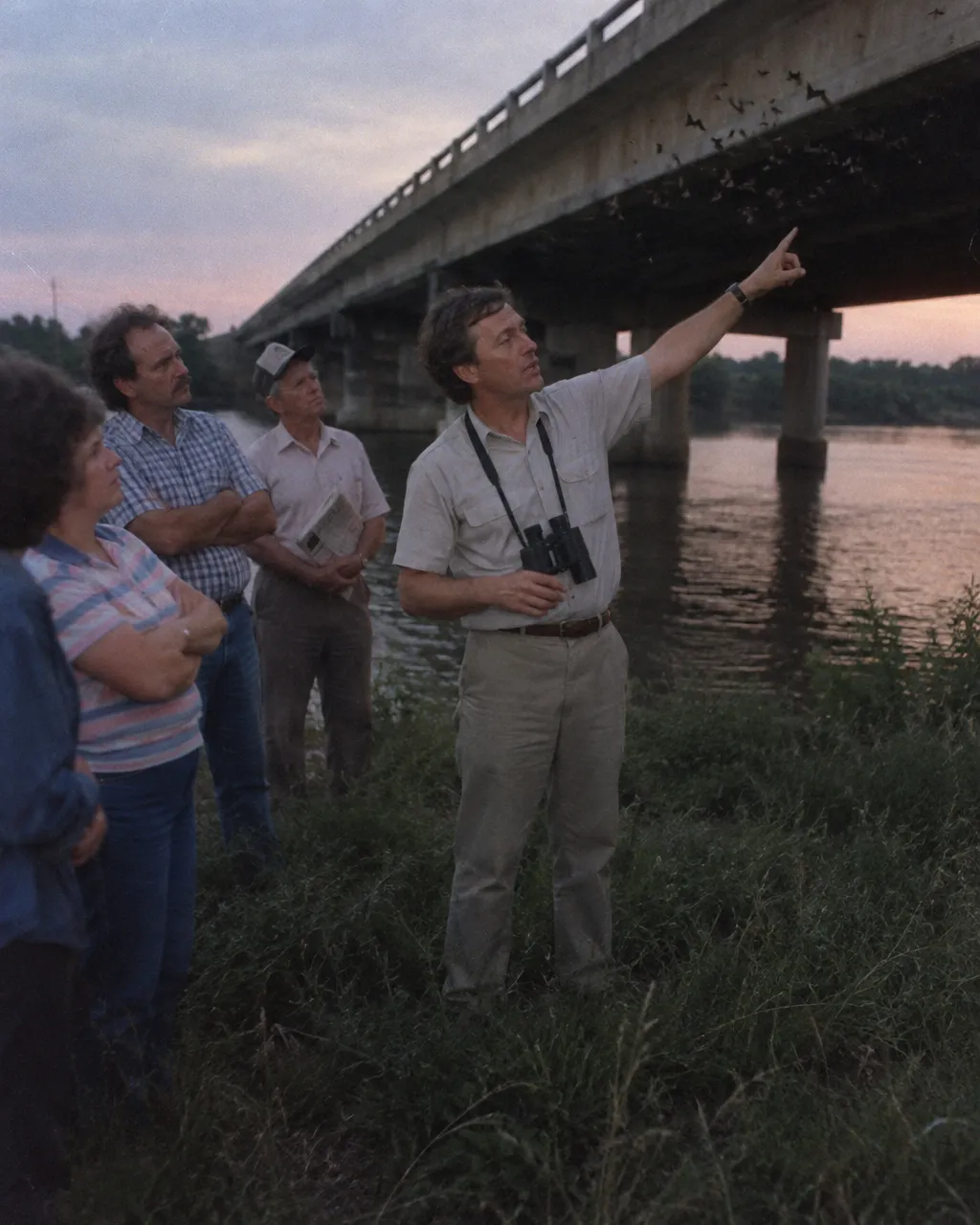 Hombre con camisa de manga corta, pantalón caqui y binoculares al cuello señala hacia la parte inferior de un puente desde la orilla de un río urbano al atardecer, en una foto con aspecto de los años ochenta.