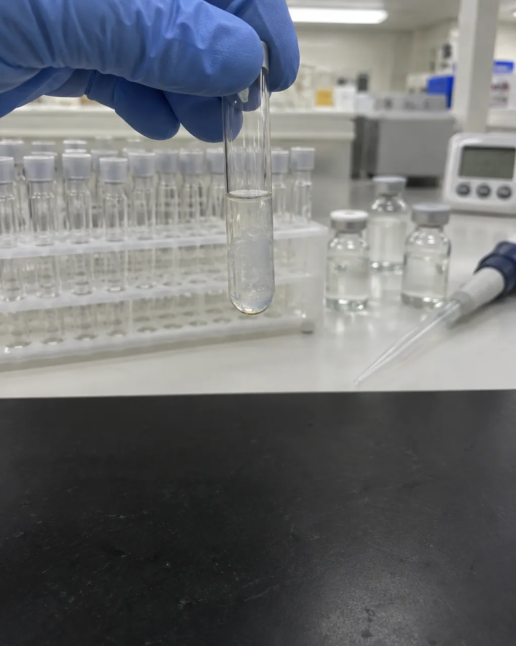 Mano con guante que sostiene boca abajo un tubo de ensayo con un gel translúcido que no se desliza, bajo la luz fluorescente de un laboratorio farmacéutico